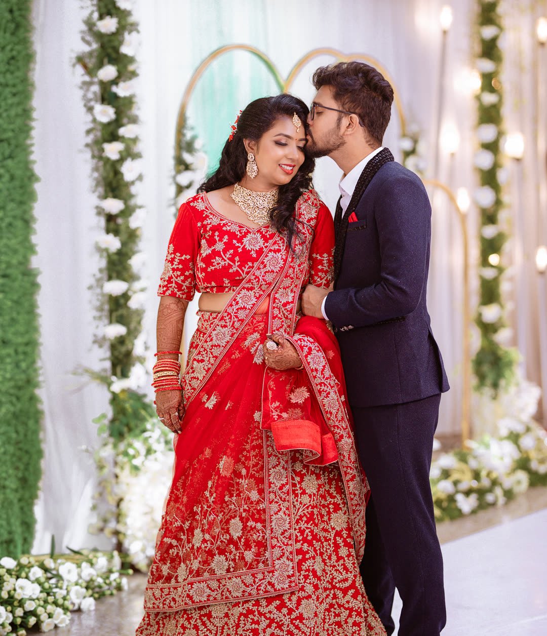 Bride in a red lehenga as the groom kisses her forehead at their reception